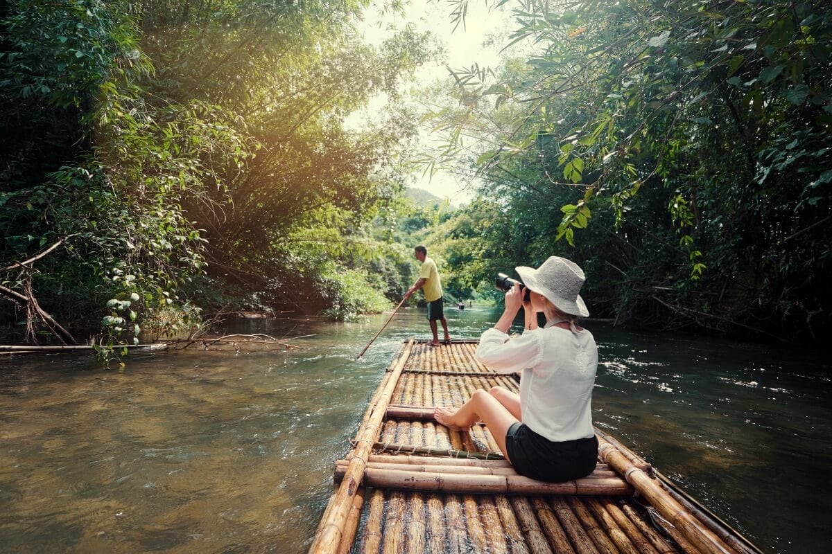 A person in a hat sits on a bamboo raft, photographing the surroundings, while a guide steers the raft along a forested river.