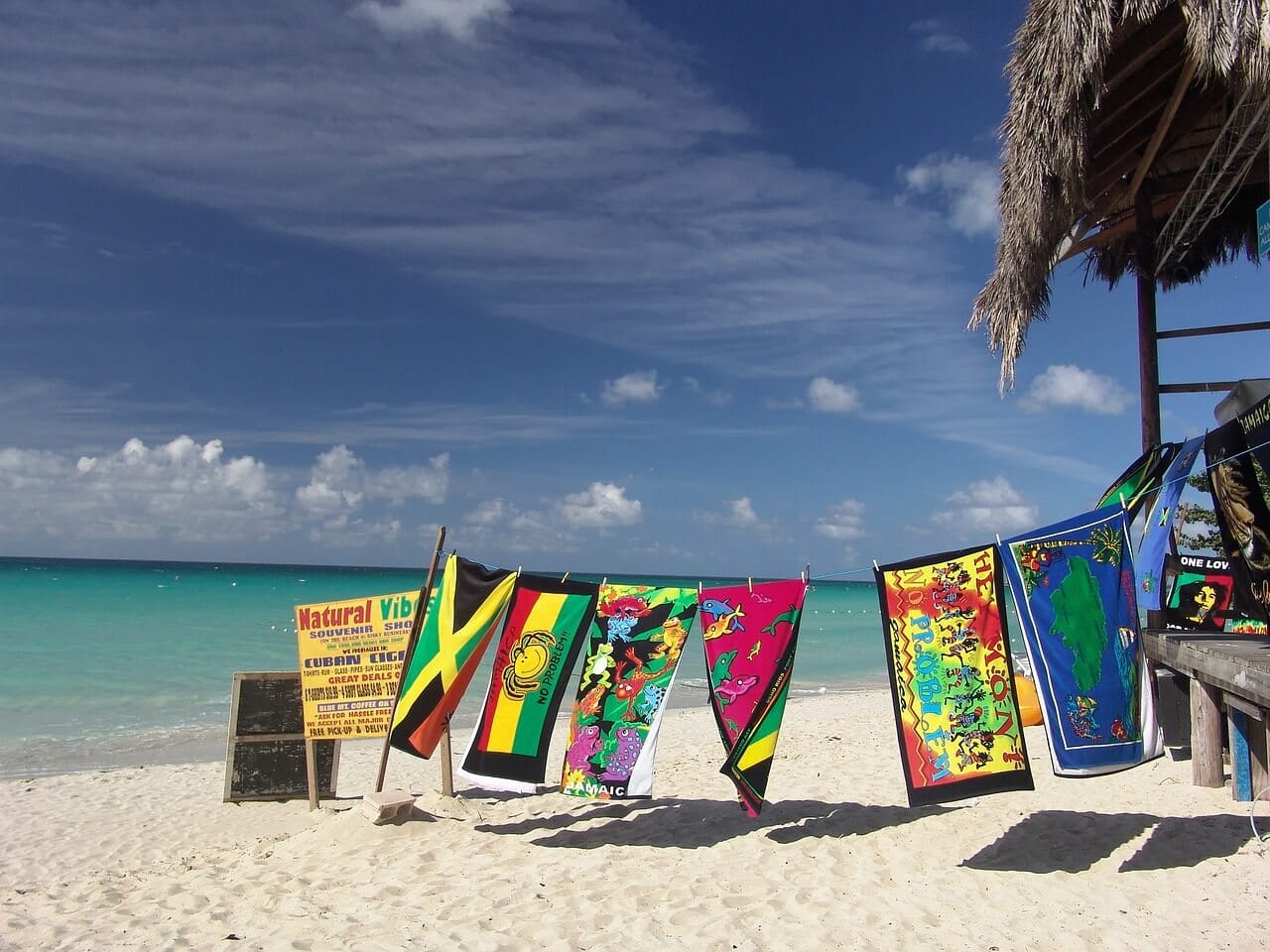 Colorful tapestries and flags hang next to a thatched hut on a sandy beach with turquoise water and a clear blue sky in the background.