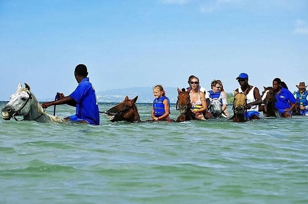 A group of people in blue life vests riding horses through waist-deep water on a sunny day.