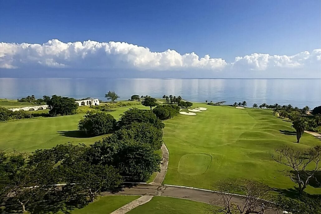 An aerial view of a lush, green golf course by a calm, blue ocean under a partly cloudy sky, with a white building to the left and sand bunkers near the water.