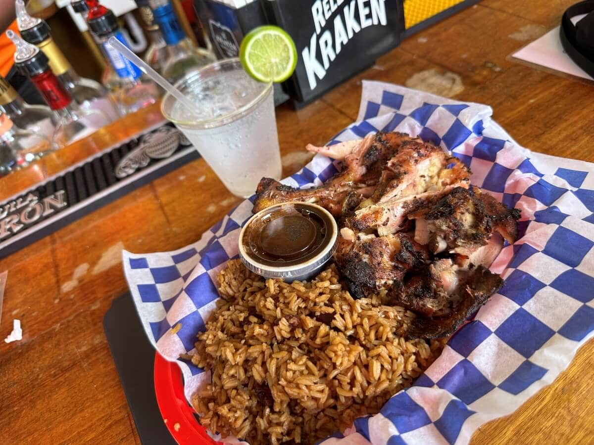 A basket with grilled chicken pieces, seasoned rice, and a small cup of sauce. A glass of a clear drink with a lime wedge is placed behind the basket, with various bottles visible in the background.