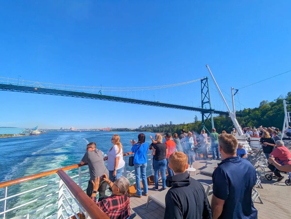 People on the Koningsdam ferry deck observing a suspension bridge and a distant city skyline under a clear blue sky during their Alaska trip.