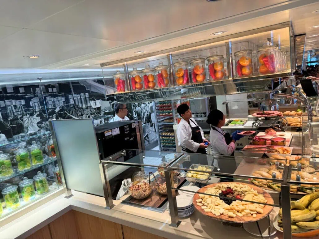 Buffet counter on Holland America's Koningsdam with chefs serving, featuring jars of pickles, fresh fruits, and assorted breads under bright lighting.