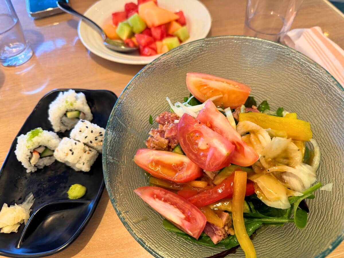 A fresh vegetable salad with tomatoes, bell peppers, and mushrooms, accompanied by sushi rolls and a side plate of fruit, served on a wooden table aboard the Koningsdam.