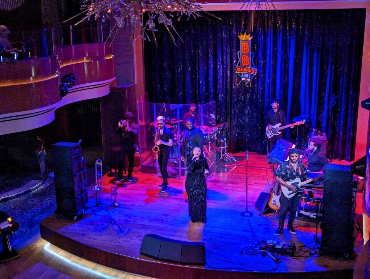 A live band performing on stage aboard the Koningsdam during a Holland America Alaska trip, featuring a vocalist, guitarists, a drummer, and horn players, with a vibrant, colorful lighting setup.