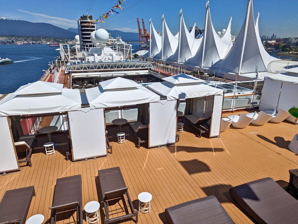 An outdoor cruise ship deck on the Holland America's Koningsdam, featuring white lounge chairs and cabanas with a view of white, sail-like structures and an Alaskan city waterfront in the background.