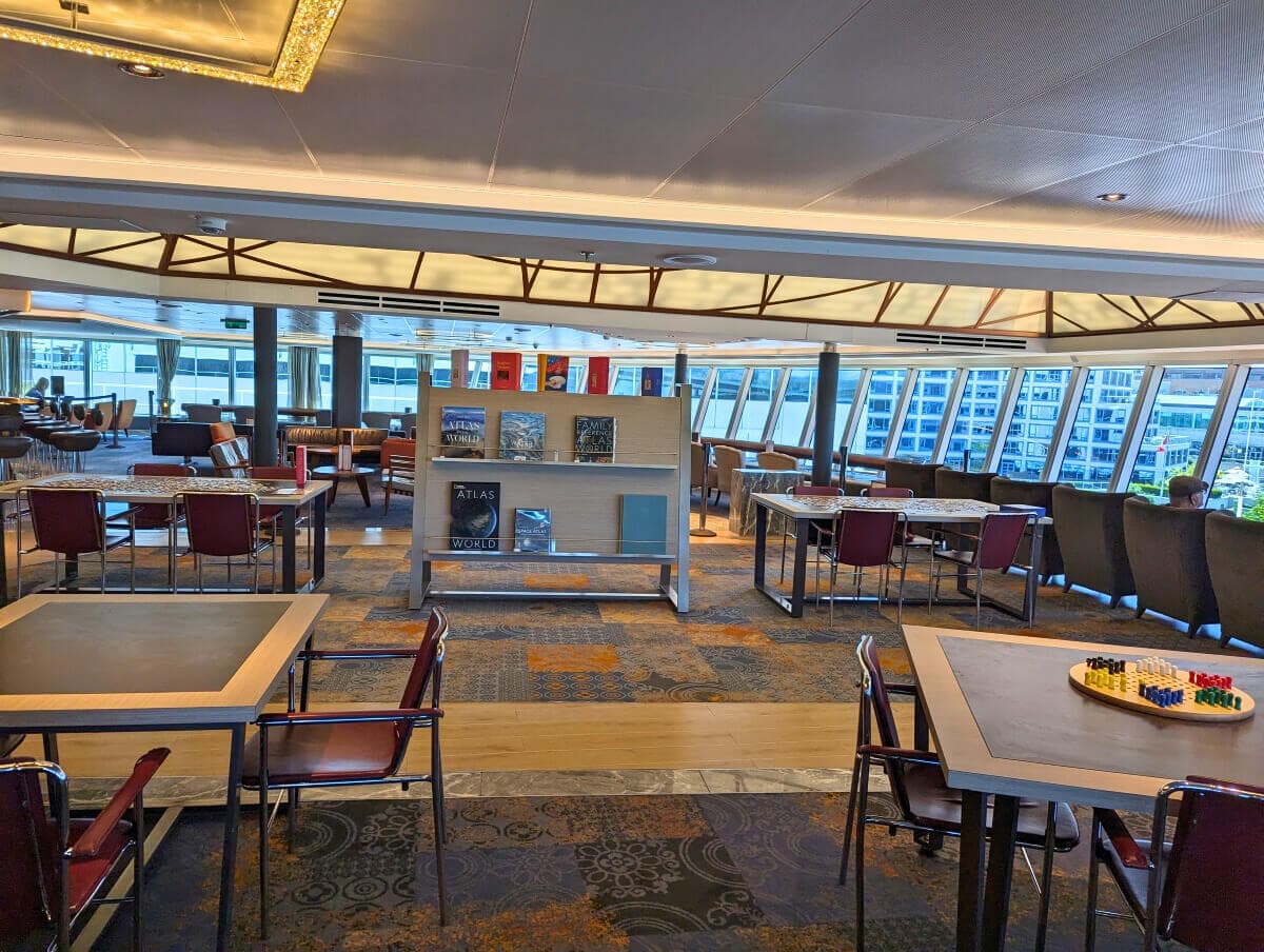 Interior of a modern library on the Koningsdam, featuring seating areas, shelves with books, and large windows overlooking city buildings.