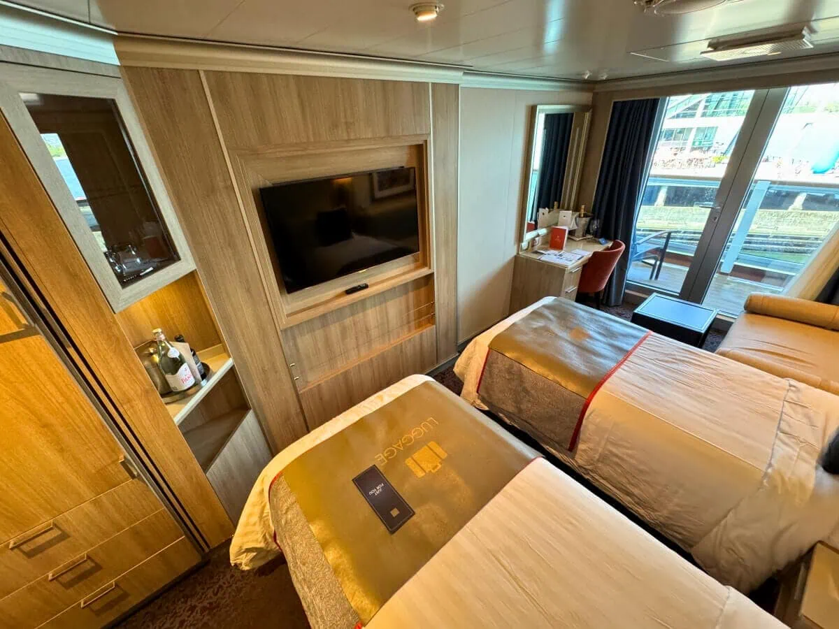 Interior of a Holland America cruise ship cabin with two twin beds, a flat-screen TV, a vanity area, and a window overlooking the harbor.