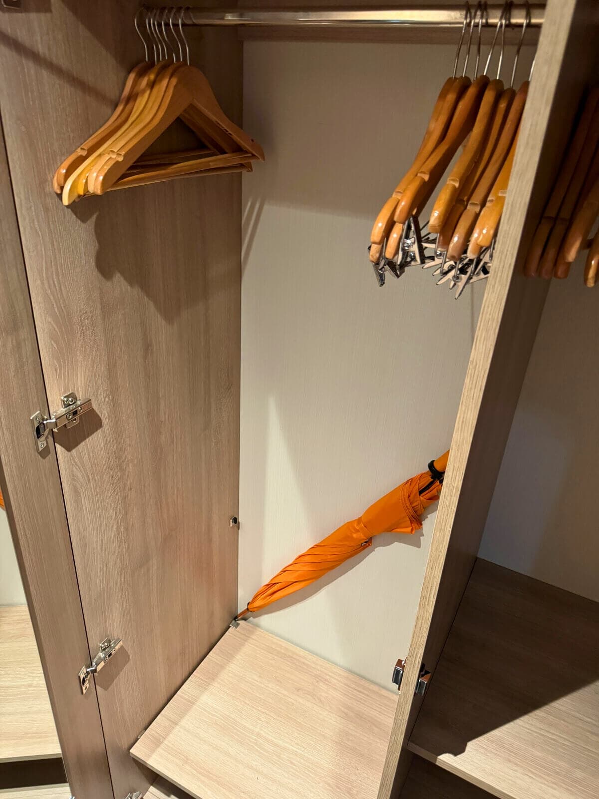 An open wooden wardrobe with empty hangers on the left side and a single orange umbrella lying on the bottom shelf, reminiscent of items one might prepare for an Alaska Trip.