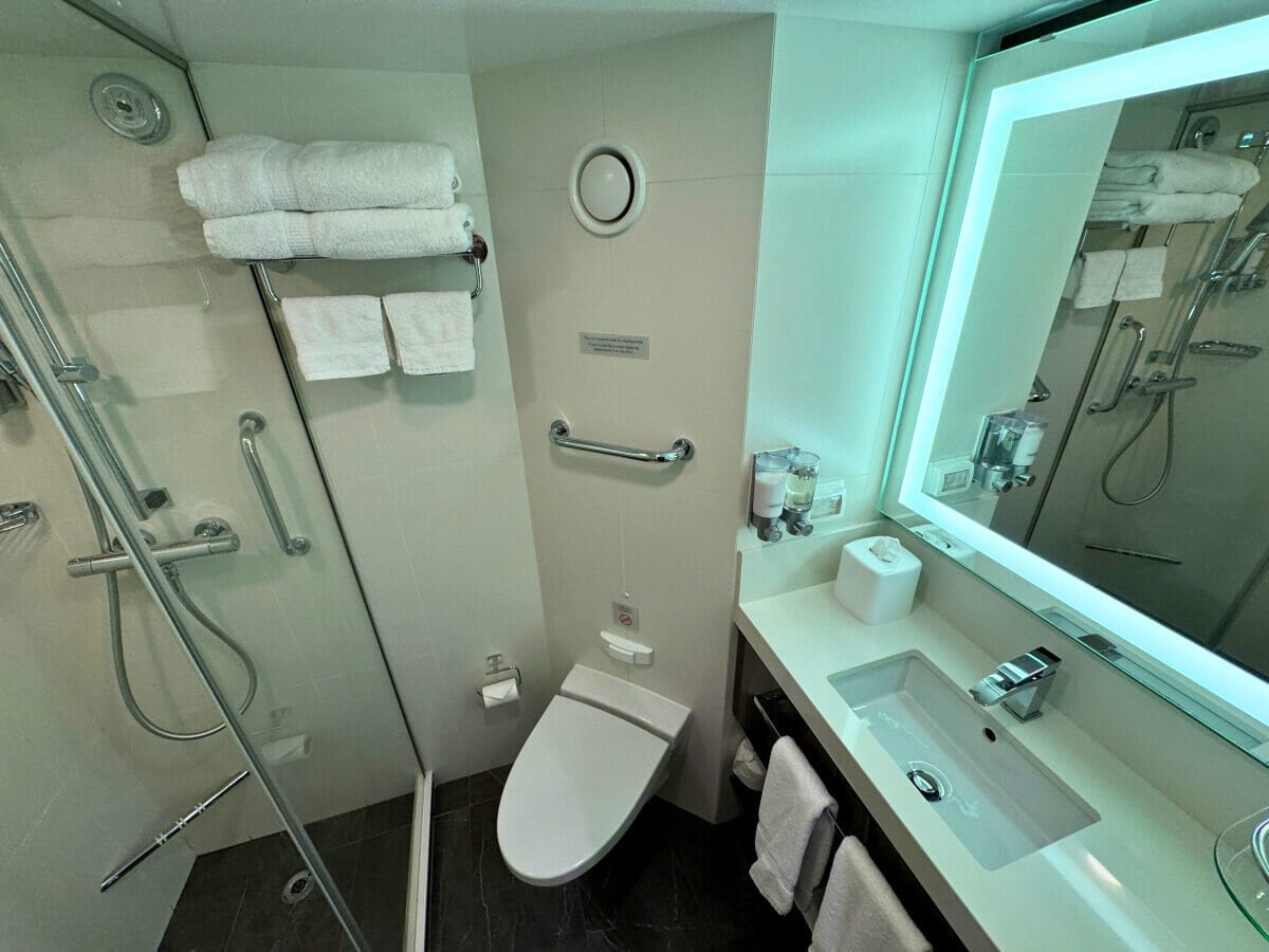 Modern bathroom interior featuring a toilet, shower stall, towel rack, sink, and mirror on Holland America's Koningsdam, with bright lighting and clean, white fixtures.