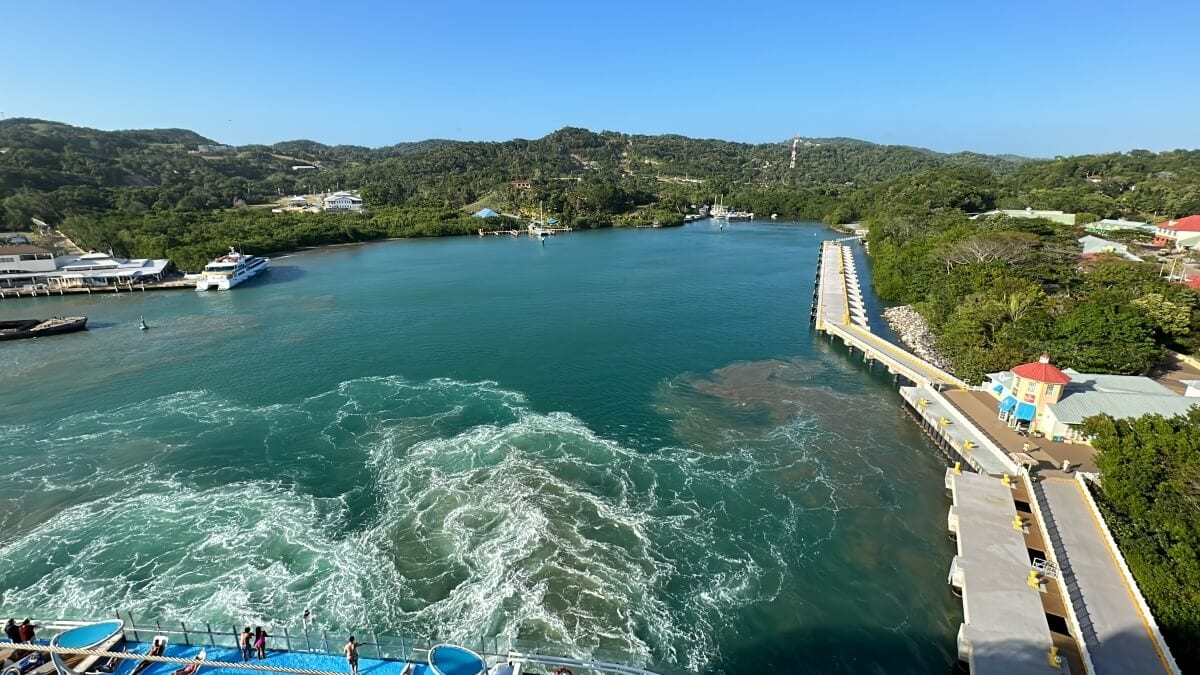 Wide aerial view of a lush green harbor with boats docked along the shores and a clear blue sky above.