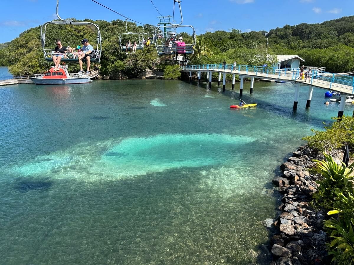 People ride a chairlift over clear blue water with visible submerged structures. A rocky shore and green foliage line the water. A bridge extends across the water next to a small boat and a kayak.