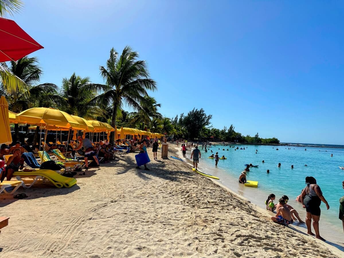 A sunny beach scene with people lounging under yellow umbrellas, swimming, and playing in the blue ocean water. Palms are scattered along the shore.