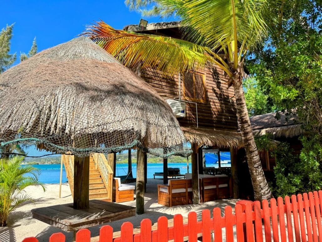 A two-story wooden hut with thatched roofs, surrounded by palm trees and a red picket fence, is situated on a sandy beach near a body of water.