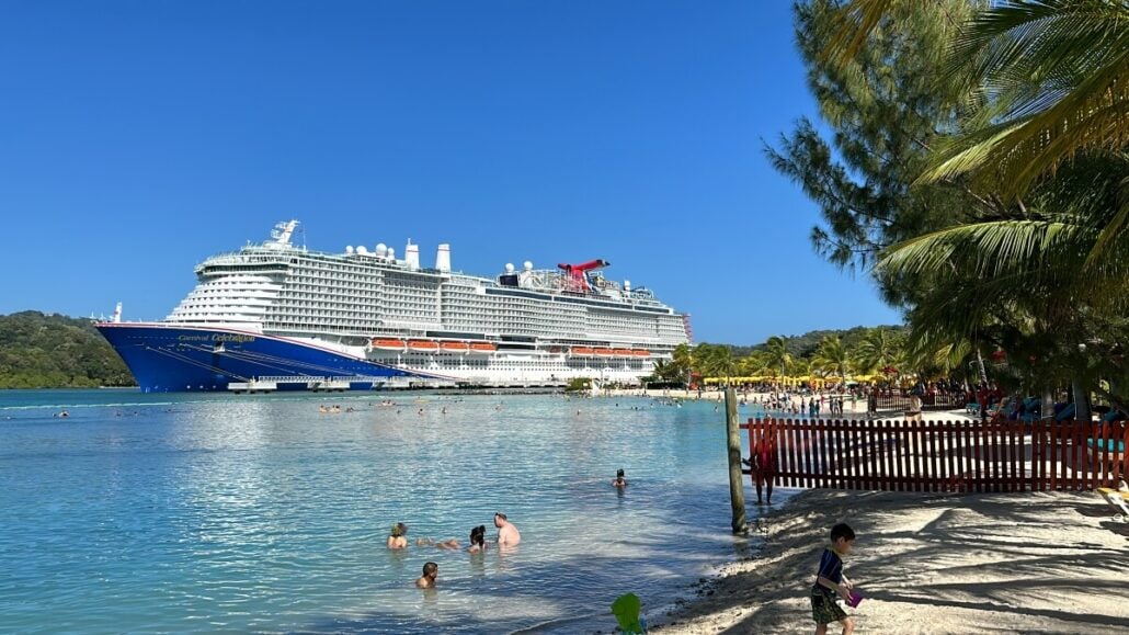 A large cruise ship is docked near a tropical beach. People are swimming in the water and relaxing on the sandy shore under clear blue skies. Palm trees border the beach.