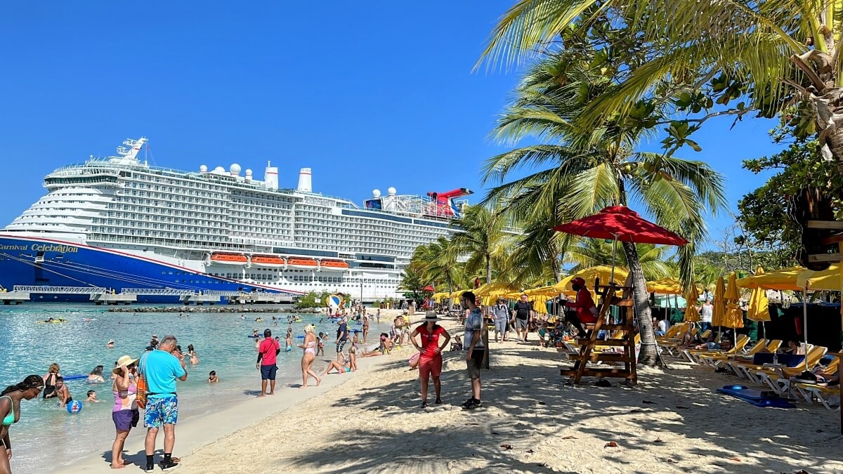 Beachgoers enjoying a sunny day at a tropical beach with palm trees, while a large cruise ship is docked in the background.