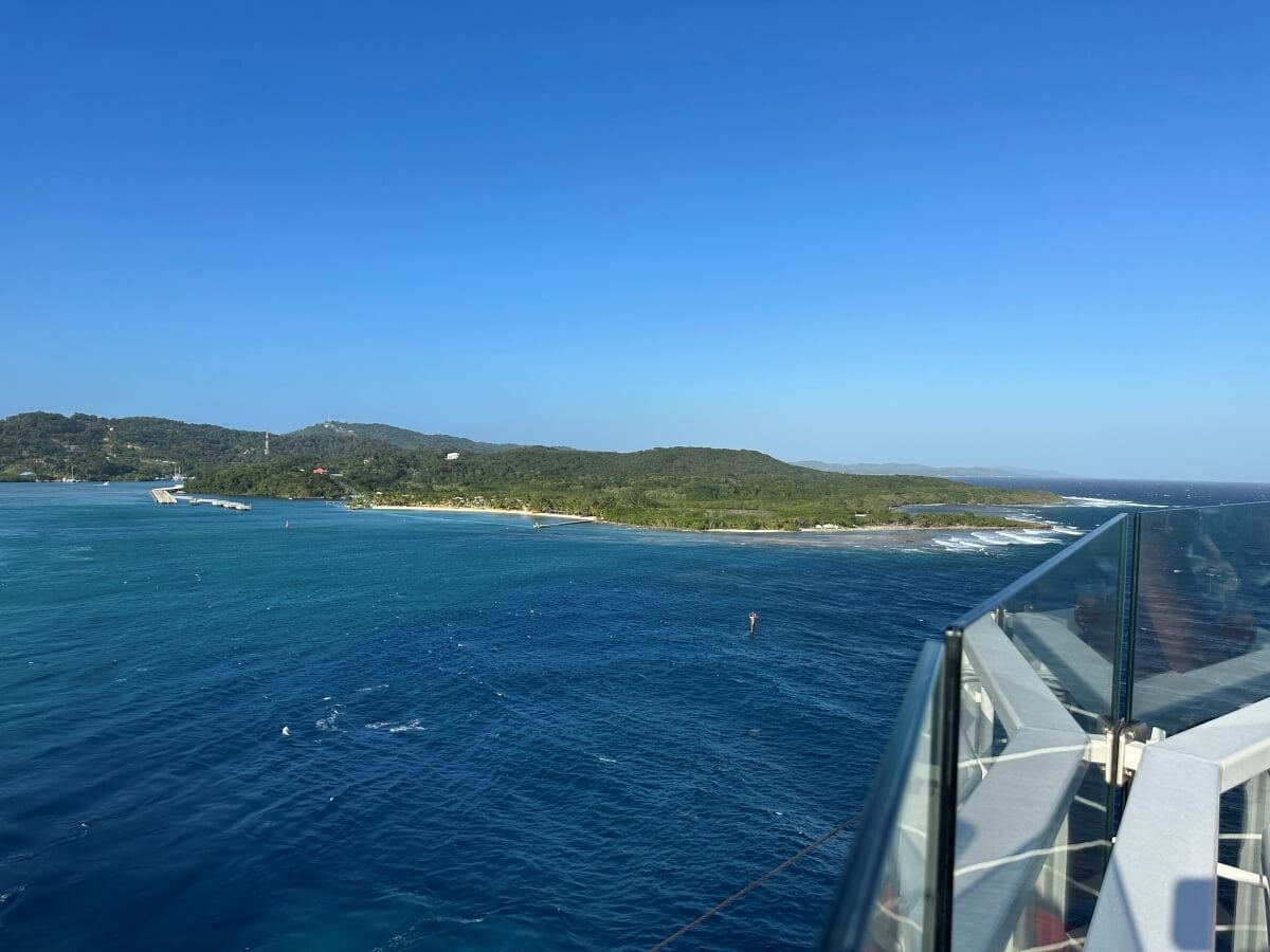 A view of a coastal landscape with clear blue ocean waters, a tree-covered shoreline, and distant hills under a clear sky, seen from a high vantage point behind a glass railing.