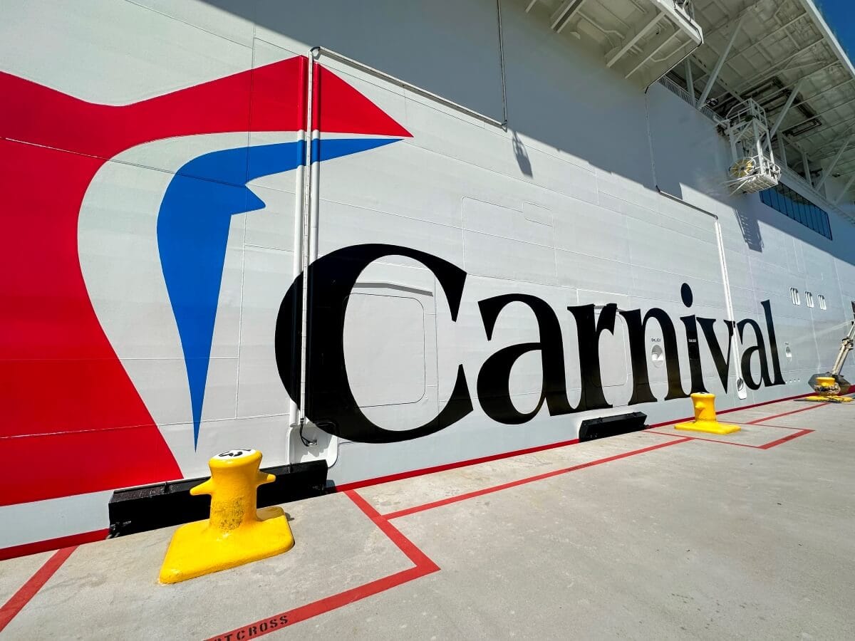 Side view of a cruise ship displaying the Carnival logo in large letters, docked at a port. Yellow bollards and red lines are visible on the concrete.