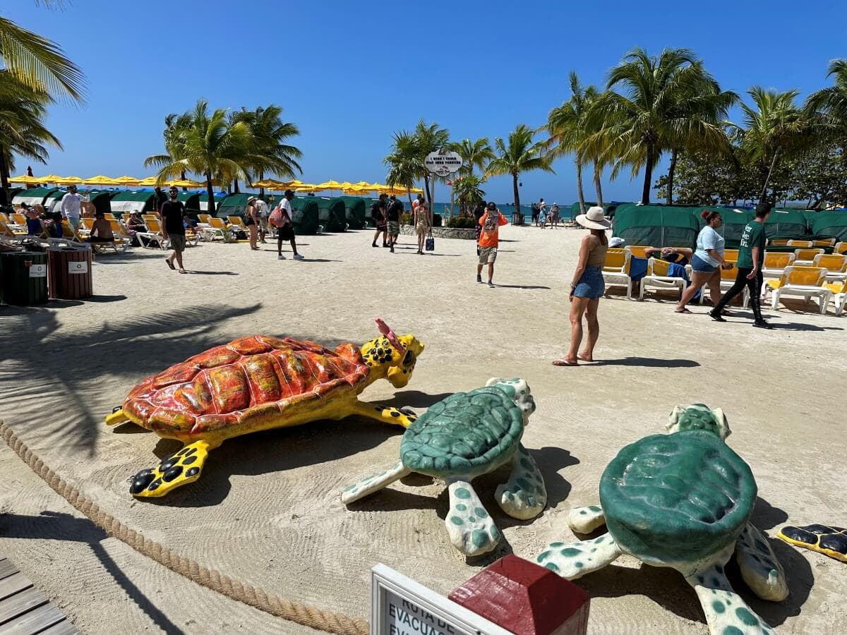 A sunny beach scene features people walking and lounging around. Three large, decorative turtle sculptures are in the foreground, amidst palm trees and beach chairs with yellow umbrellas in the background.