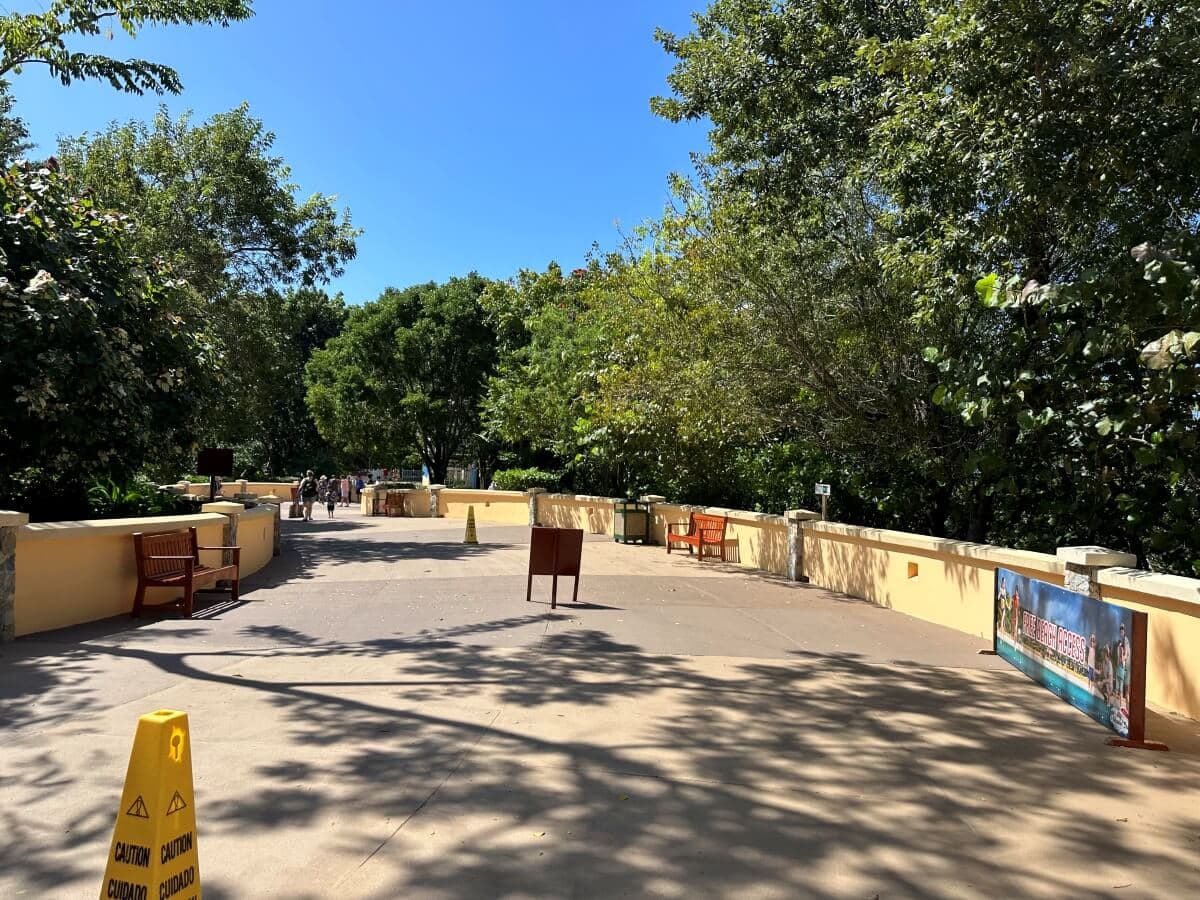 A wide paved walkway is bordered by leafy trees and yellow railings. Red benches and a sign are visible along the path. Yellow caution signs stand on the walkway. The sky is clear and blue.