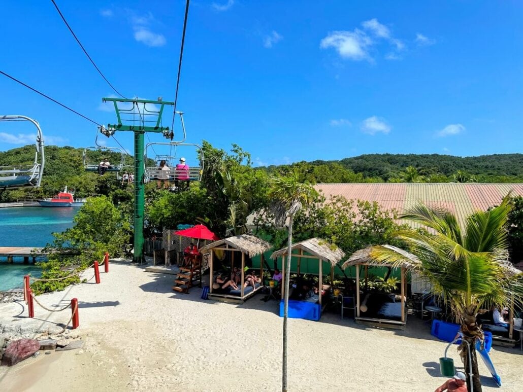 A beach scene with a sandy area, several thatched-roof shelters, and people relaxing on lounge chairs. A cable car system runs overhead near the shoreline with lush greenery and a blue sky in the background.