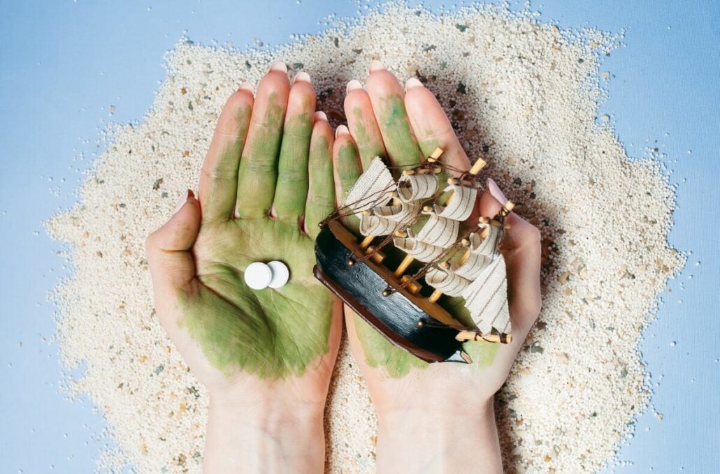 Hands with green paint hold a miniature wooden ship and two white pills, surrounded by sand on a blue surface, hinting at remedies for seasickness on a cruise.