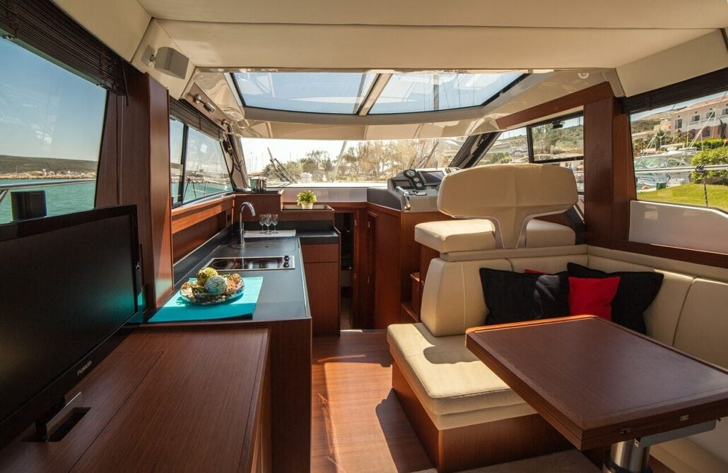 Interior view of a boat’s cabin featuring a kitchen area with a stove and sink, a seating area with a table and cushions, and windows offering a scenic view outside. Discover the convenience for those looking to rent a boat at an affordable price.