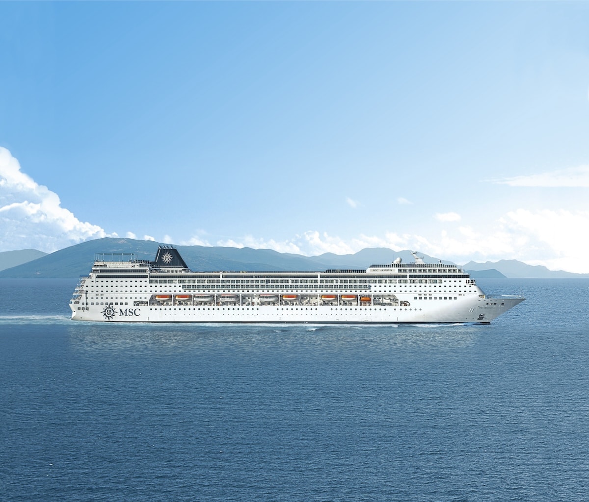 A large MSC cruise ship sailing on a clear blue ocean with mountains in the distant background under a sunny sky near the coast of Spain.