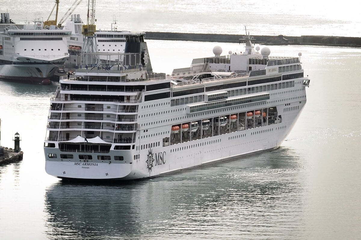 A large white cruise ship named MSC Armonia, an elegant vessel from MSC Cruises, is docked at a port, with a second cruise ship visible in the background.