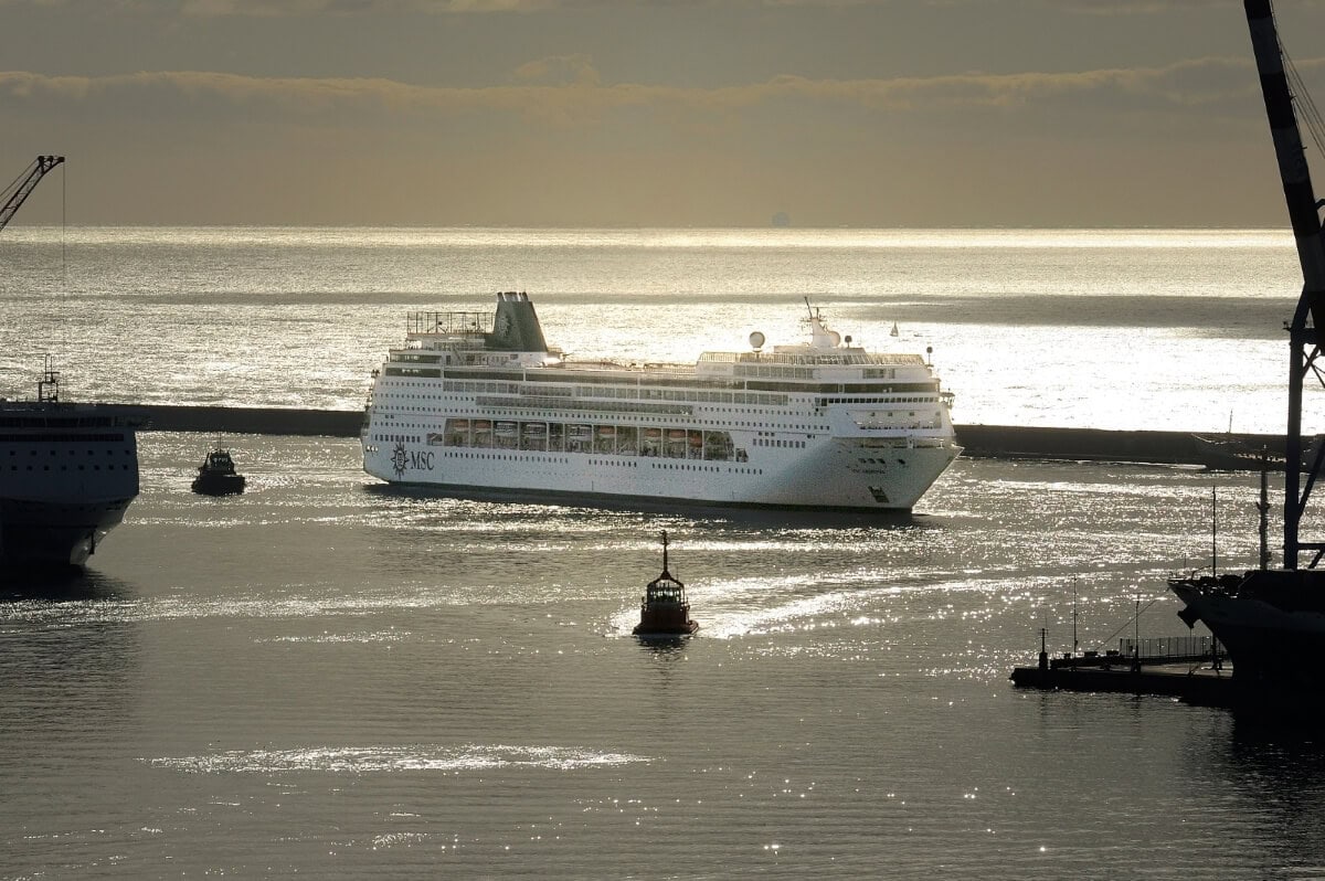 An MSC Cruises ship sails on a sunlit sea toward a harbor with smaller boats nearby and silhouetted cranes in the background.