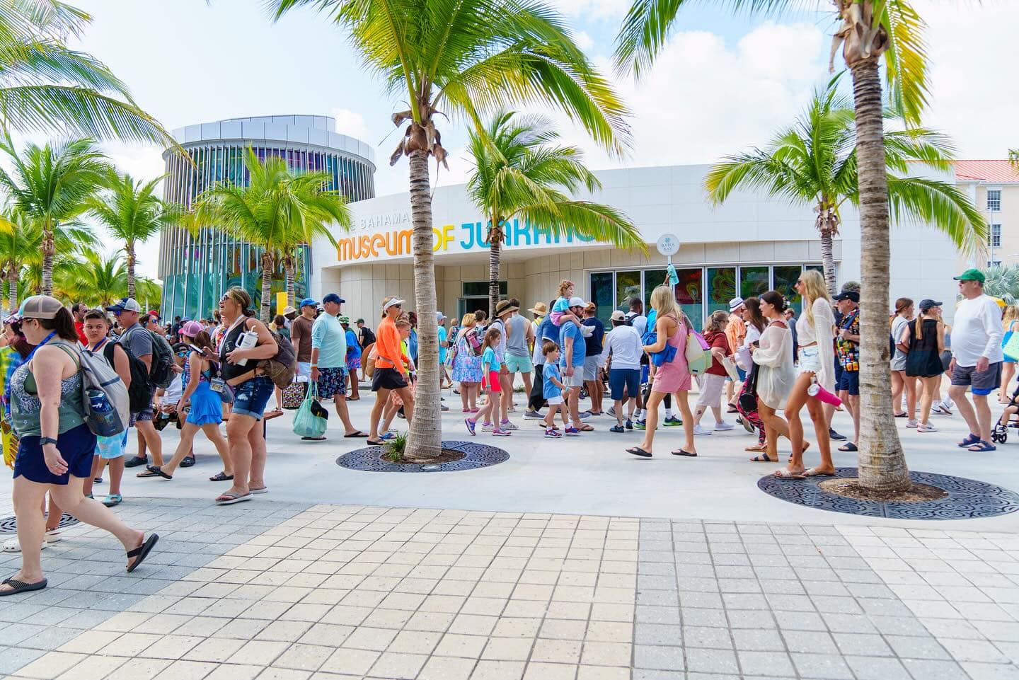 A large crowd of people walking outside the Museum of Junkanoo, set amidst palm trees near the Nassau Cruise Port on a bright, sunny day.