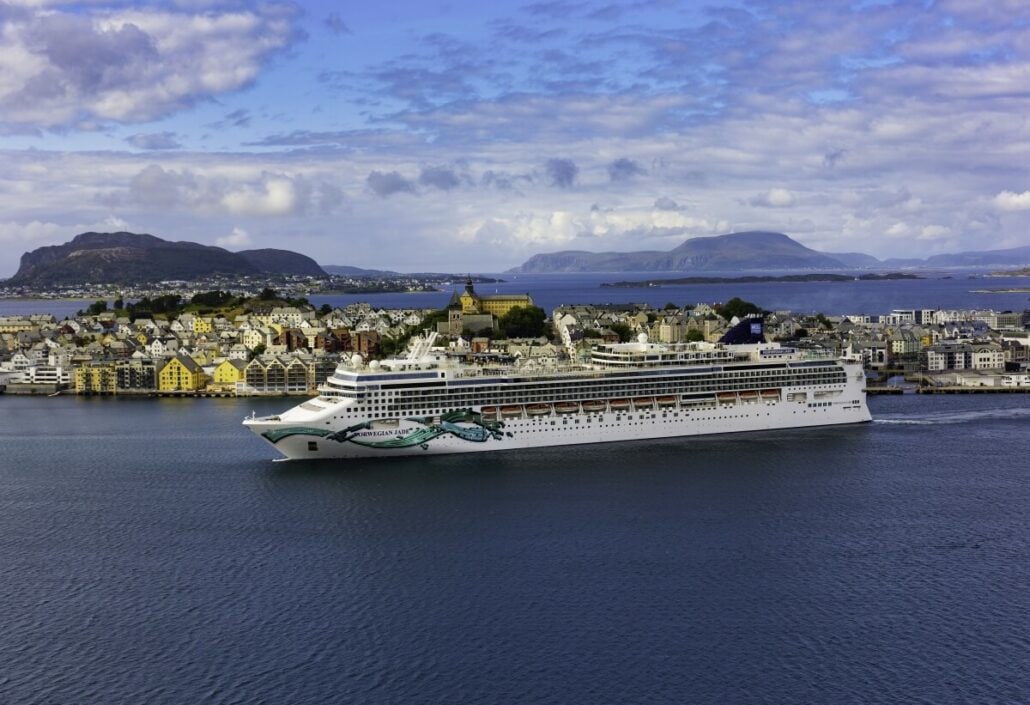 A white cruise ship, the Norwegian Jade, sails near a coastal town with colorful buildings, mountains, and a partly cloudy sky in the background.