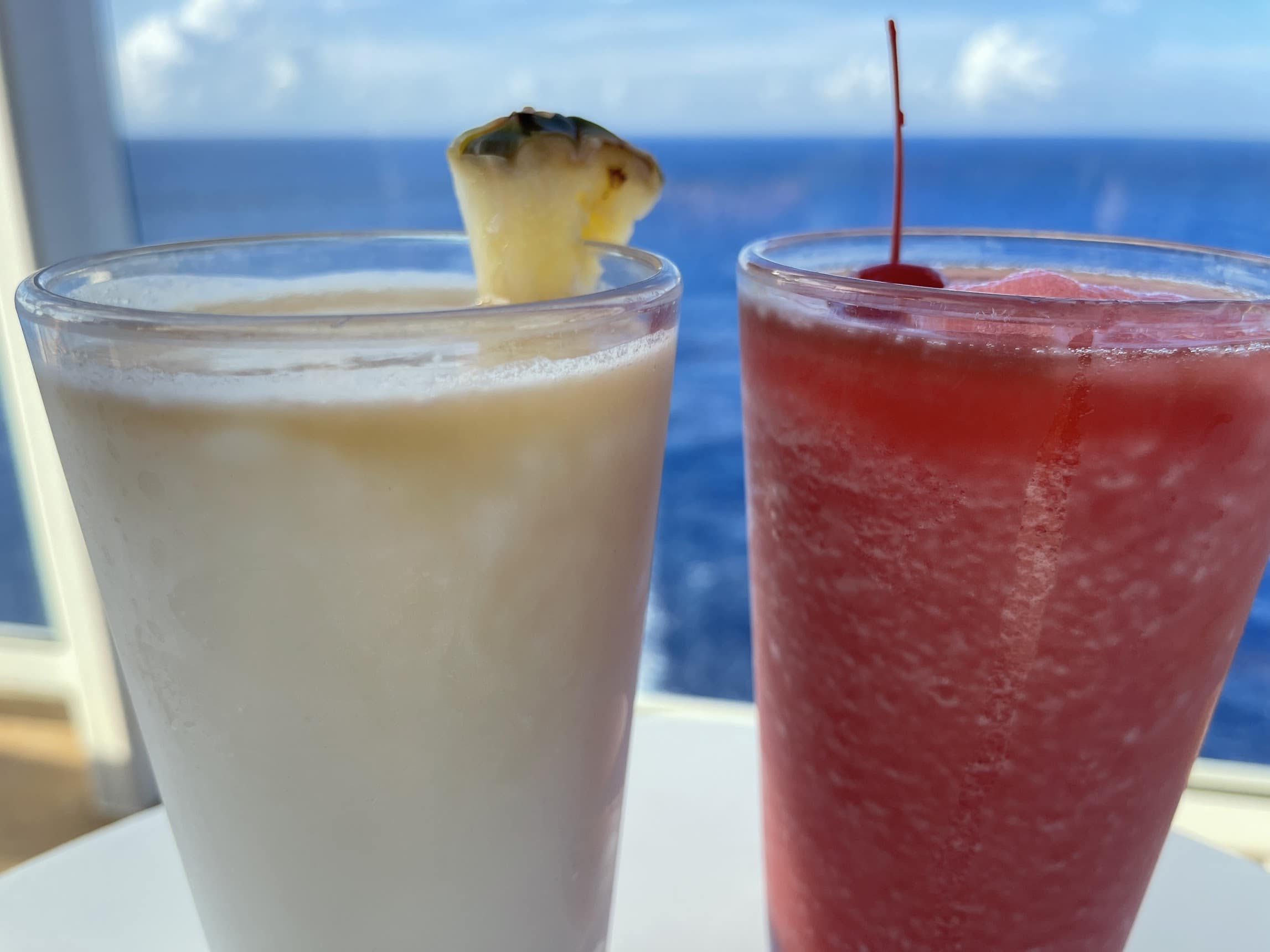 Two glasses of tropical drinks, one milky white with a pineapple garnish, the other strawberry red with a cherry, against an ocean background, perfect for passengers 18 to 20.