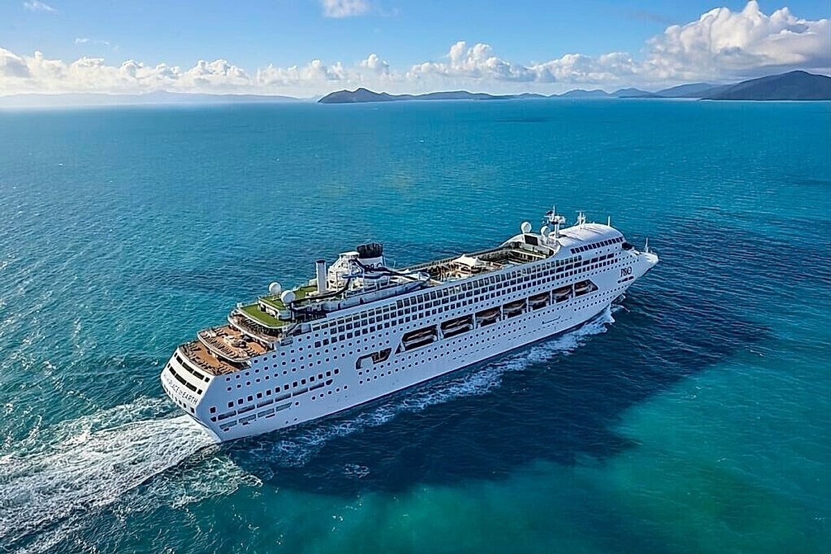 An aerial view of a large white cruise ship, one of the recently sold Carnival ships, sailing on a calm blue ocean with distant islands and a partly cloudy sky in the background.