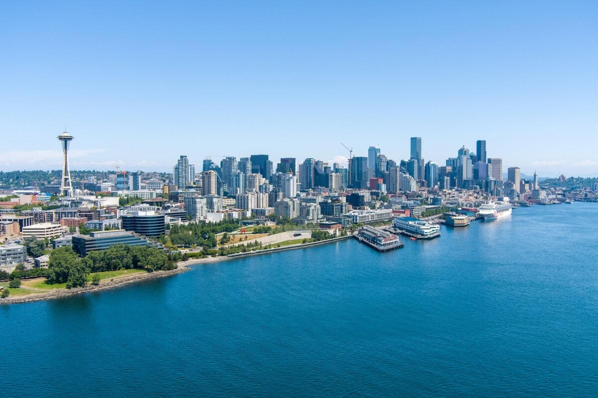 Aerial view of a city skyline with numerous high-rise buildings along the waterfront. The Space Needle is visible on the left side, and there are docks and docked cruise ships equipped with shore power in the foreground. Clear blue sky above.