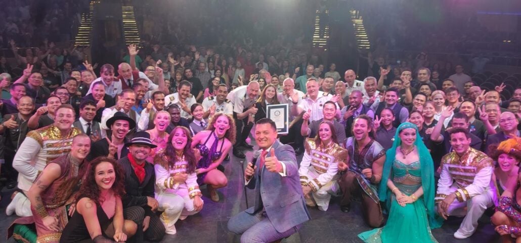 A large group of people, many in costume, pose together on a stage at the Giant Pizza Party. A person at the center holds up a framed certificate for the Guinness World Record, while another person next to them gives a thumbs up to the camera.