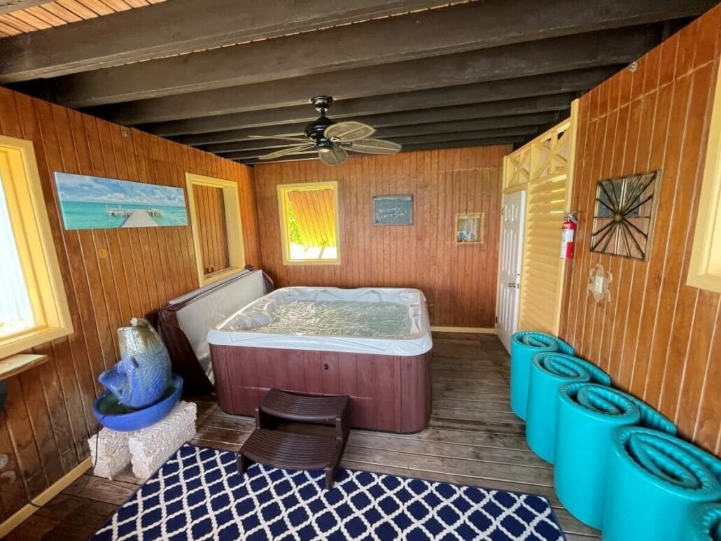 A cozy Half Moon Cay cabana with wooden walls features a hot tub, ceiling fan, wall paintings, rolled-up blue mats, a small table, and a patterned rug.