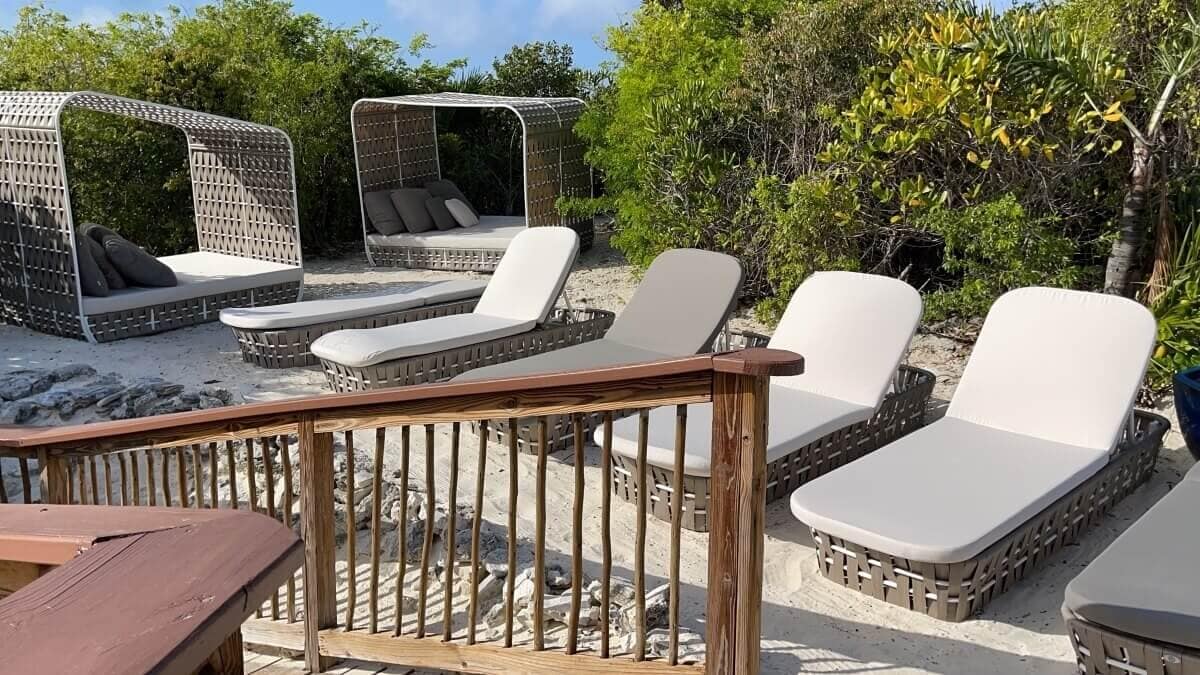 Outdoor seating area with wicker lounge chairs and Half Moon Cay cabanas on sand, surrounded by greenery. Wooden railing in the foreground.