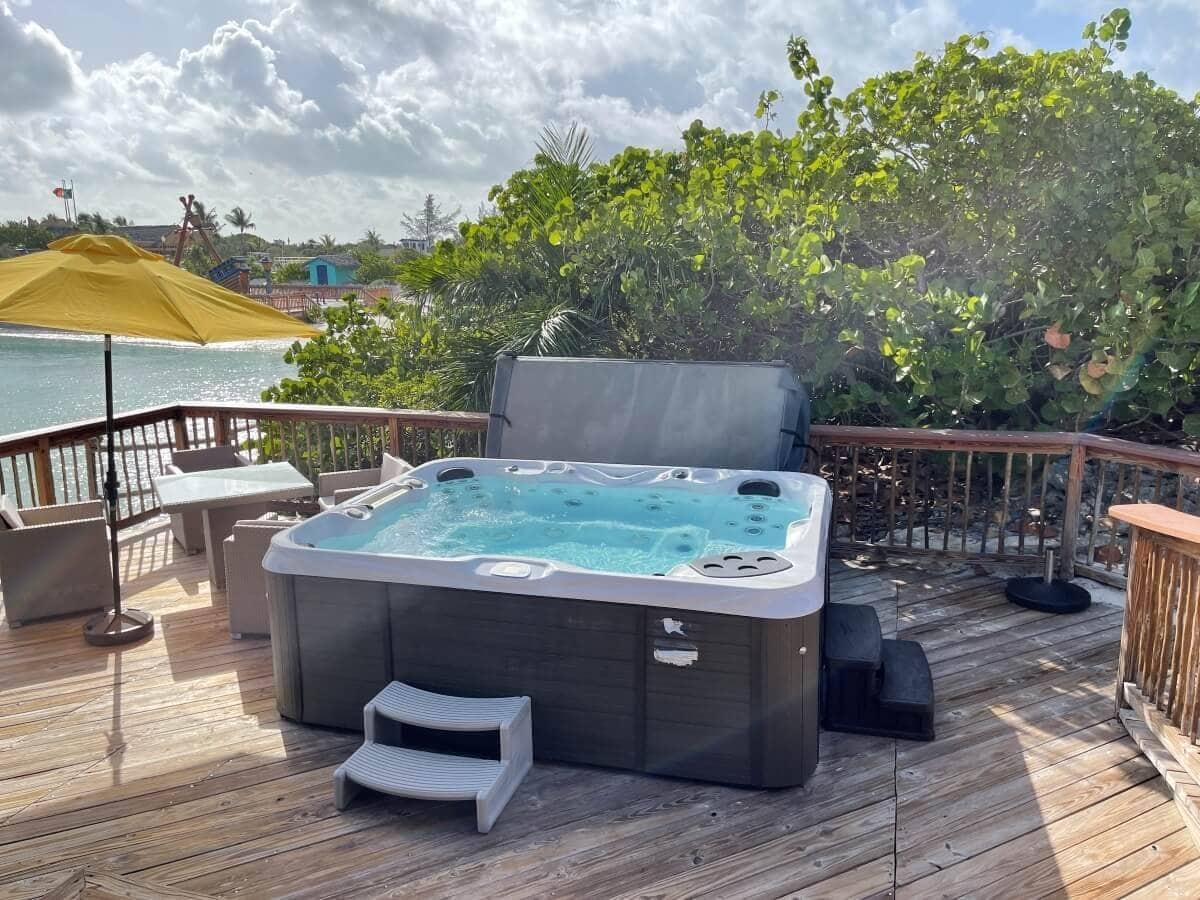 A Half Moon Cay cabana features a hot tub on a wooden deck overlooking a body of water. There's an umbrella, chairs, and lush greenery in the background.