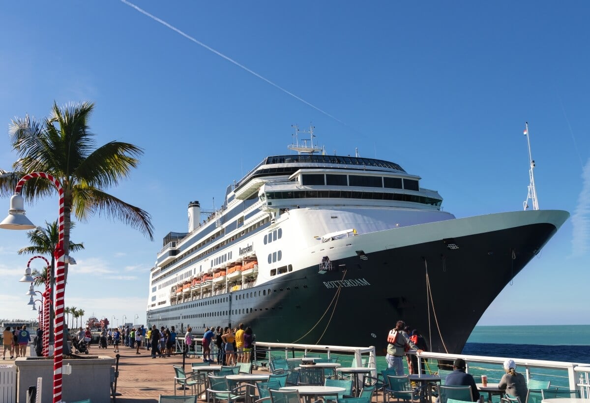 A large cruise ship docked at a pier on a sunny day with people walking around and palm trees nearby.