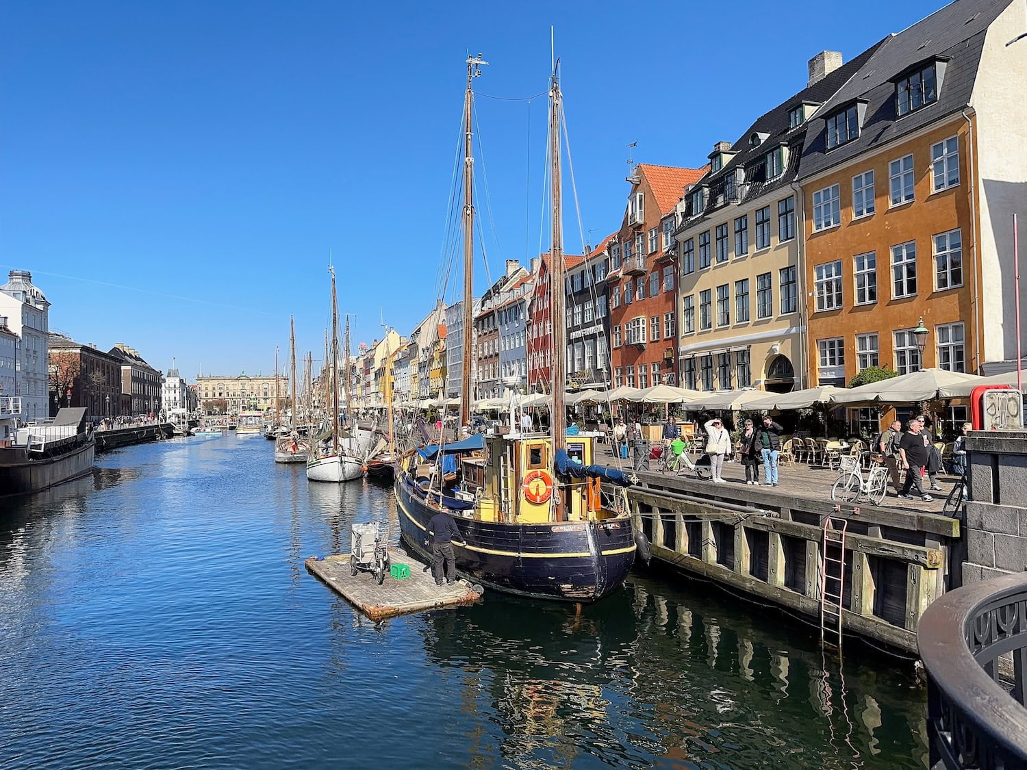 A canal lined with colorful buildings and restaurants on the right. Moored boats are on the water, available to rent at an affordable price, with a clear blue sky above.