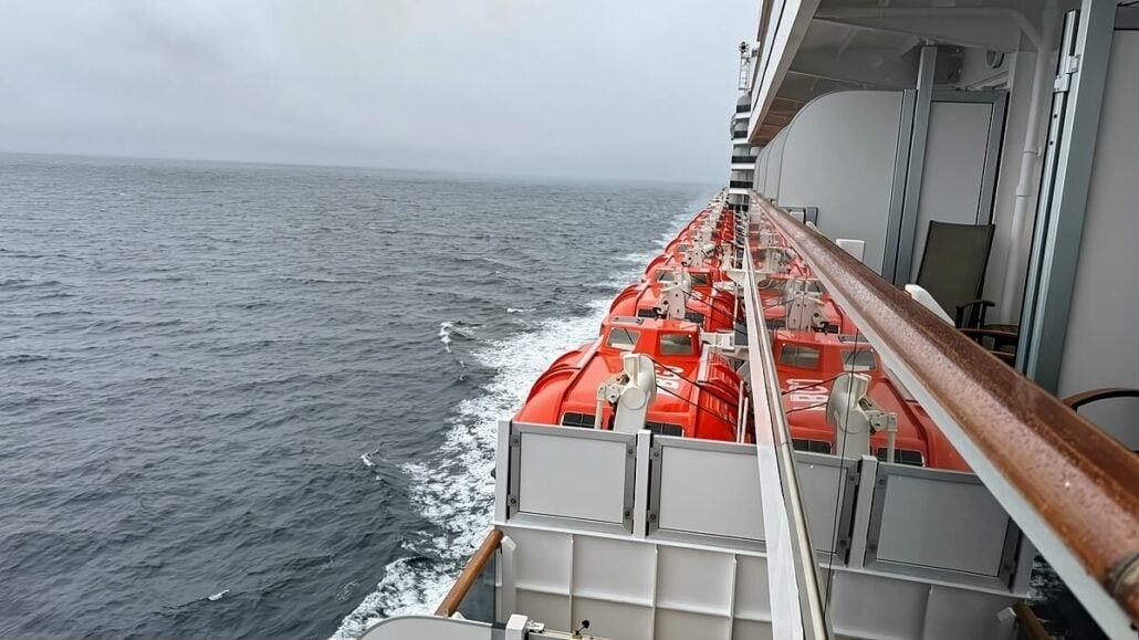 View of the ocean from the side of a cruise ship on an Alaska trip, showing a row of orange lifeboats and the scenic Inside Passage.