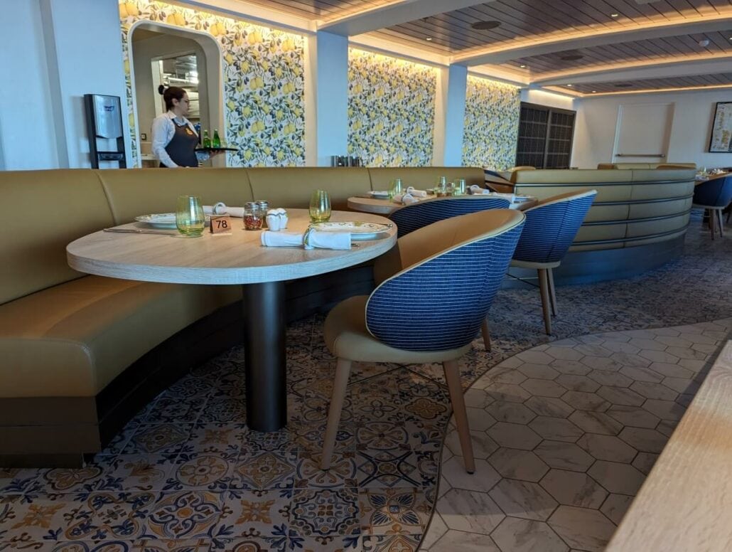 A neatly arranged restaurant interior with patterned flooring, a floral wall design, and a server in the background.