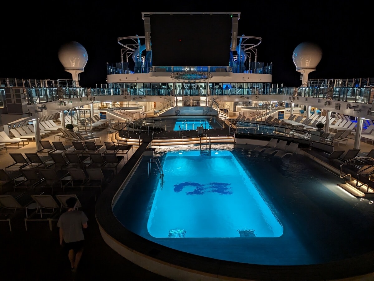 Illuminated cruise ship pool deck at night with loungers and a large screen.