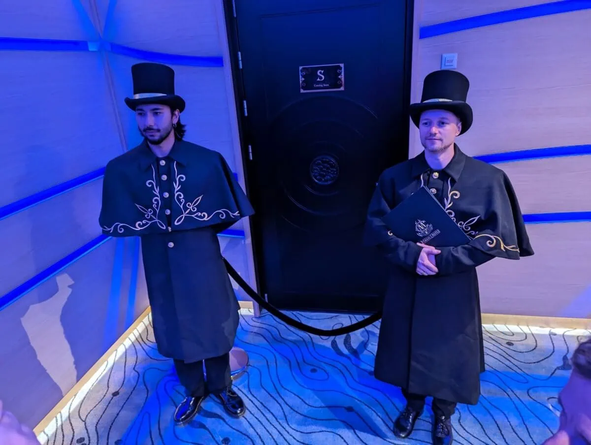 Two individuals dressed in traditional formal attire with top hats standing in a blue-lit room.