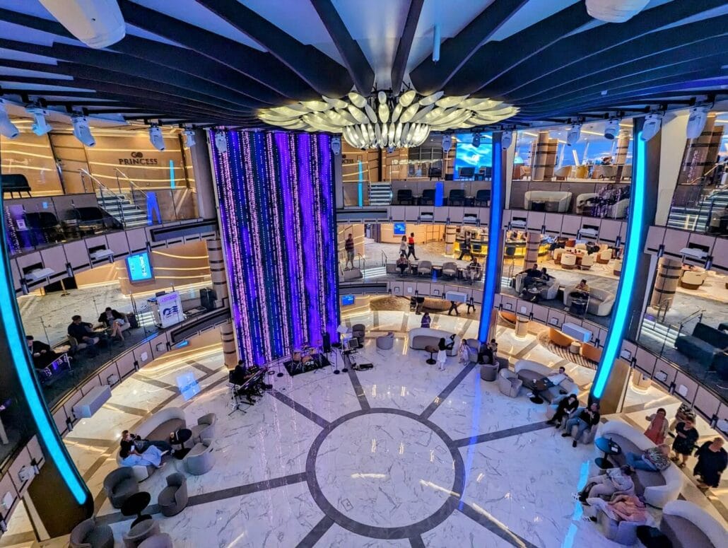 Overhead view of the Sun Princess, a modern multi-level cruise ship atrium with passengers relaxing on sofas, an illuminated purple pillar, and balconies.