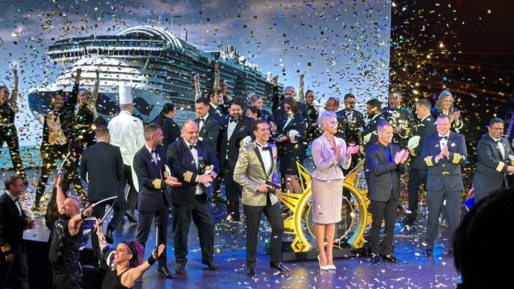 People celebrating on stage at a ship launch event, confetti in the air, with a woman turning a ship's wheel in the center.