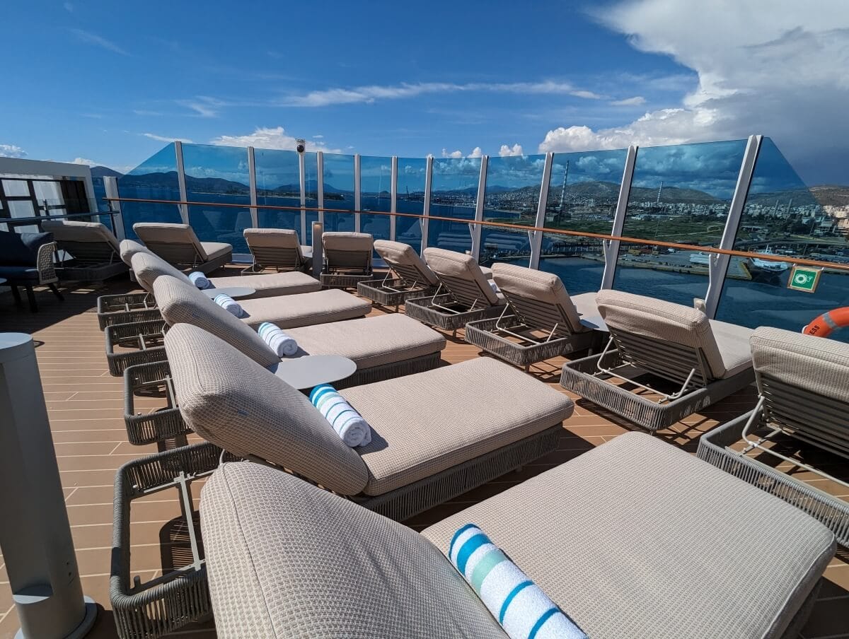 Sun loungers lined up on a cruise ship deck with a glass barrier overlooking the sea and distant landscape.