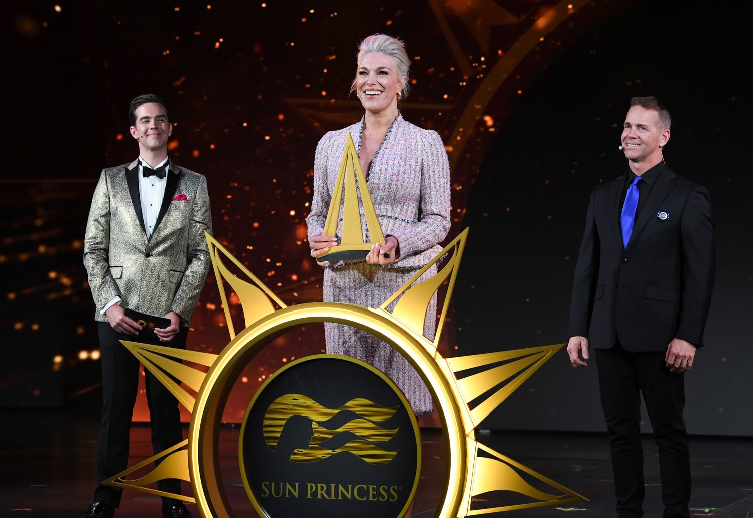 Hannah Waddingham holding an award on stage at the Sun Princess event, flanked by two men, with a festive golden backdrop.