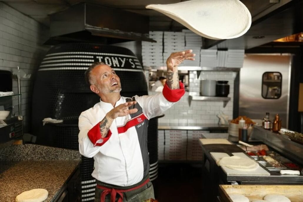 A chef, aiming for a Guinness World Record, expertly tosses pizza dough in the kitchen with a pizza oven in the background. He is sporting a white and red chef's coat, preparing for Princess Cruises' giant pizza party.